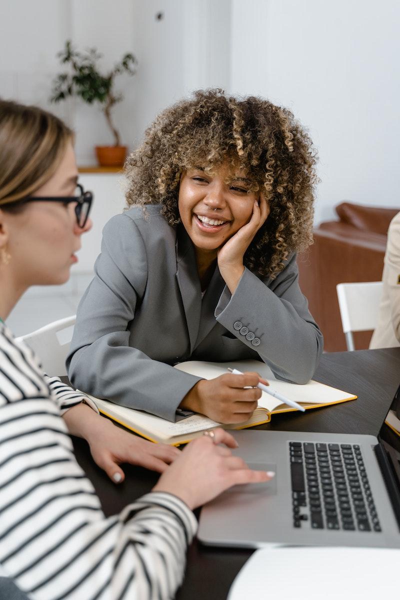 Photo by MART PRODUCTION A Woman in Gray Blazer Smiling while Looking at Her Colleague Using a Laptop